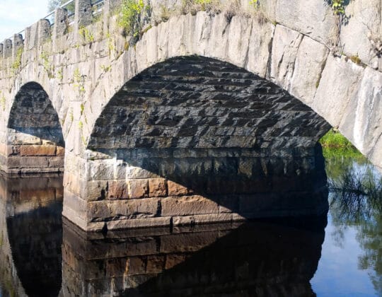 Historisk stenbro över Helge å med bågformade valv och stilla vatten som speglar konstruktionen
