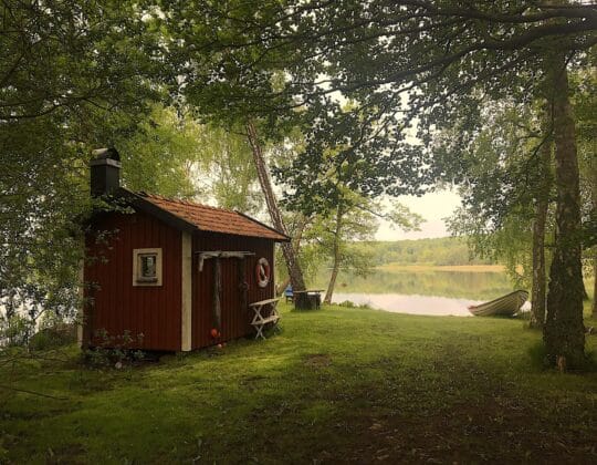 Litet rött träskjul vid en stilla sjö, omgivet av grönskande träd och en uppdragen roddbåt.